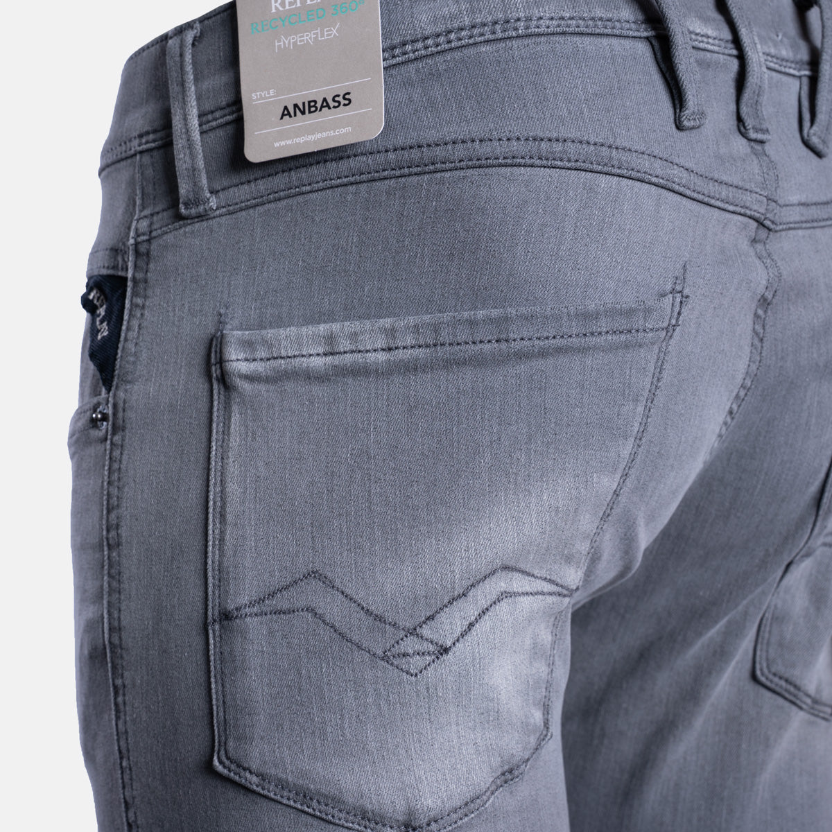Close-up of the back waistband and pocket area of gray denim jeans, showing a metal brand tag that reads ANBASS. Replay Jeans available at Sakks Menswear in Southampton.