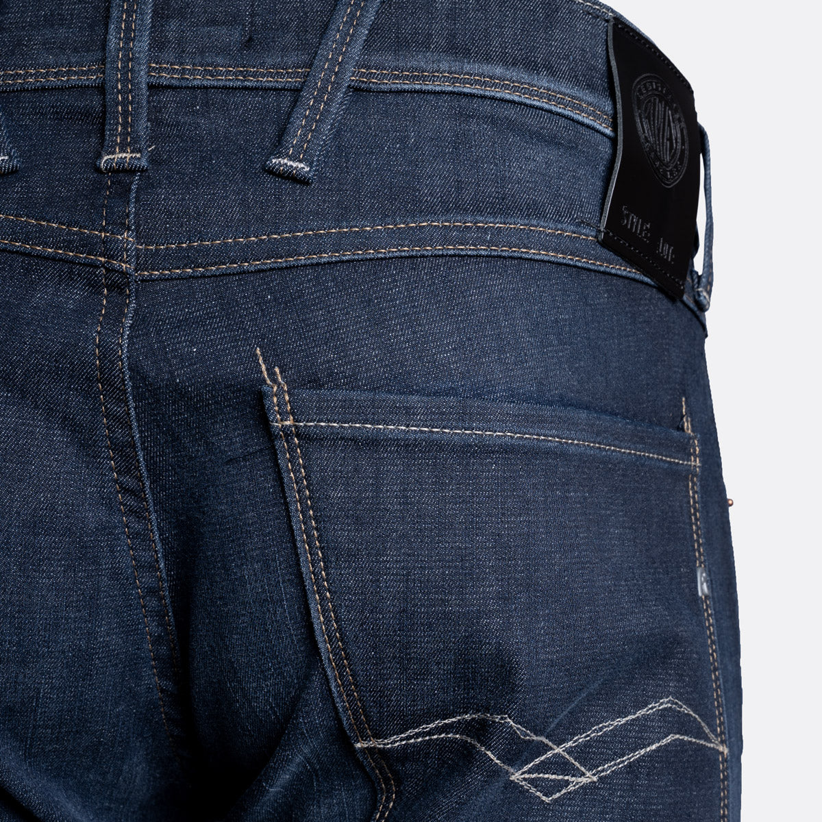 Close-up of the back waistband and pocket area of dark blue denim jeans, showing contrast stitching, belt loops, and a black brand patch on the waistband. Replay Jeans available at Sakks Menswear in Southampton.