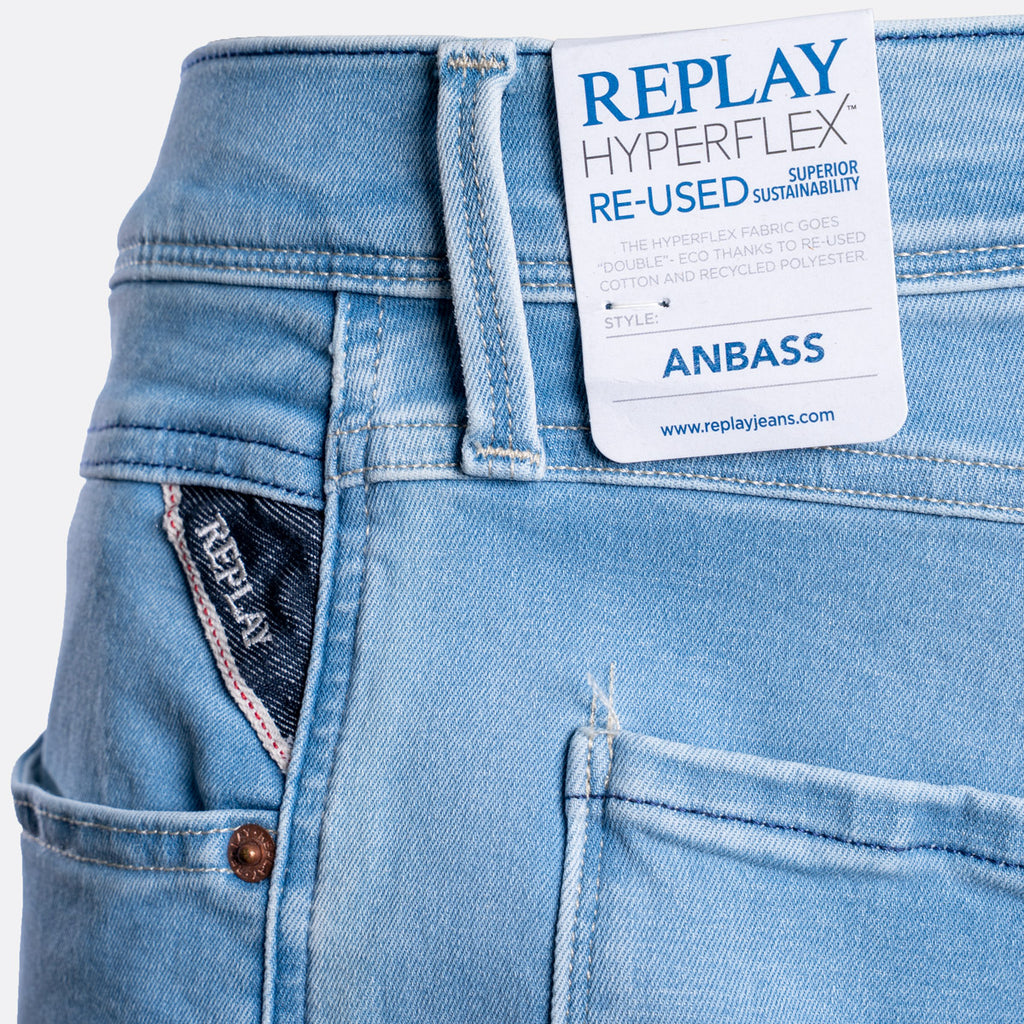 Close-up of light blue denim jeans showing the waistband, back pocket, button, and a white REPLAY paper label attached at the back waist. The label reads 'REPLAY HYPERFLEX RE-USED SUSTAINABILITY' and 'ANBASS'. Replay Jeans available at Sakks Menswear in Southampton.