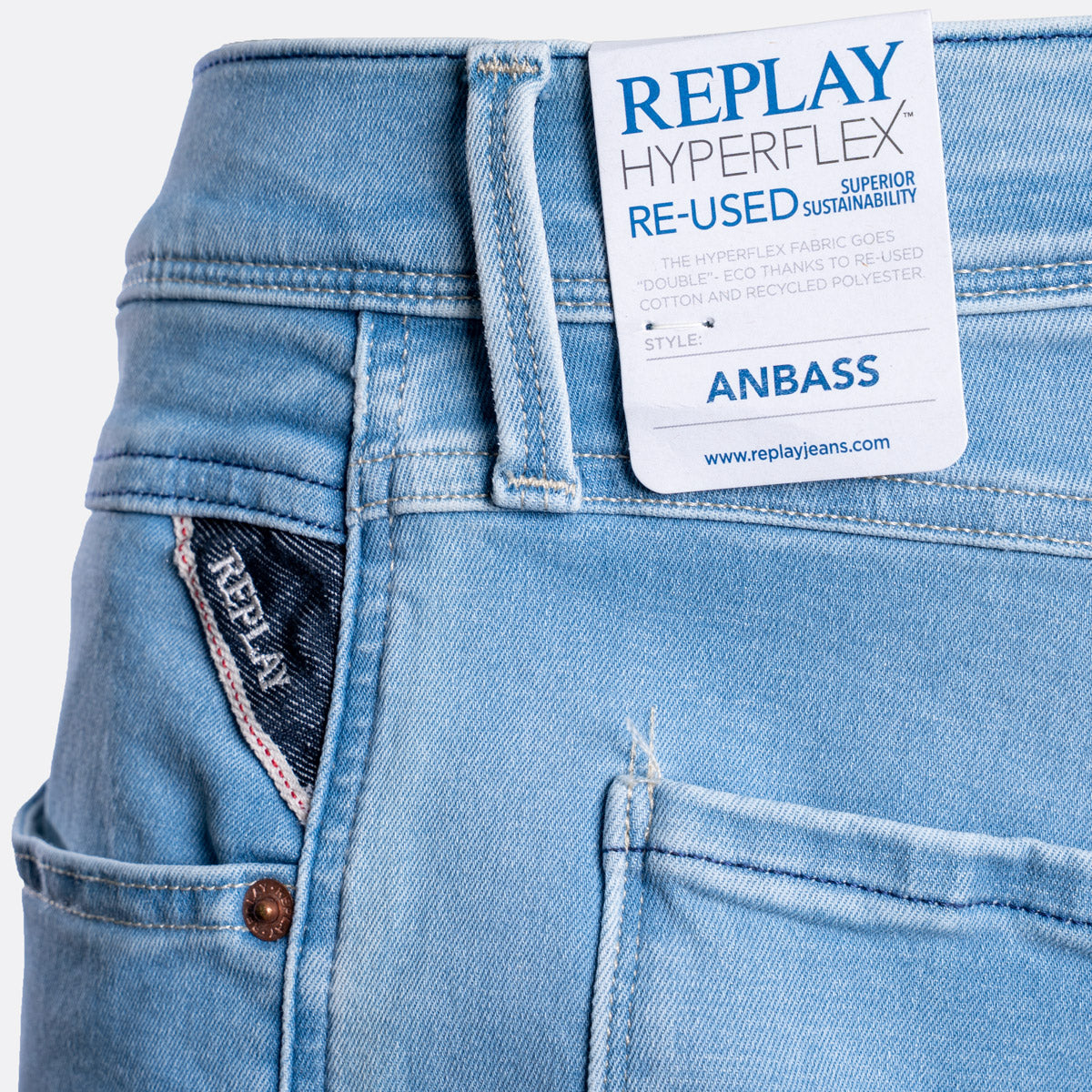 Close-up of light blue denim jeans showing the waistband, back pocket, button, and a white REPLAY paper label attached at the back waist. The label reads 'REPLAY HYPERFLEX RE-USED SUSTAINABILITY' and 'ANBASS'. Replay Jeans available at Sakks Menswear in Southampton.