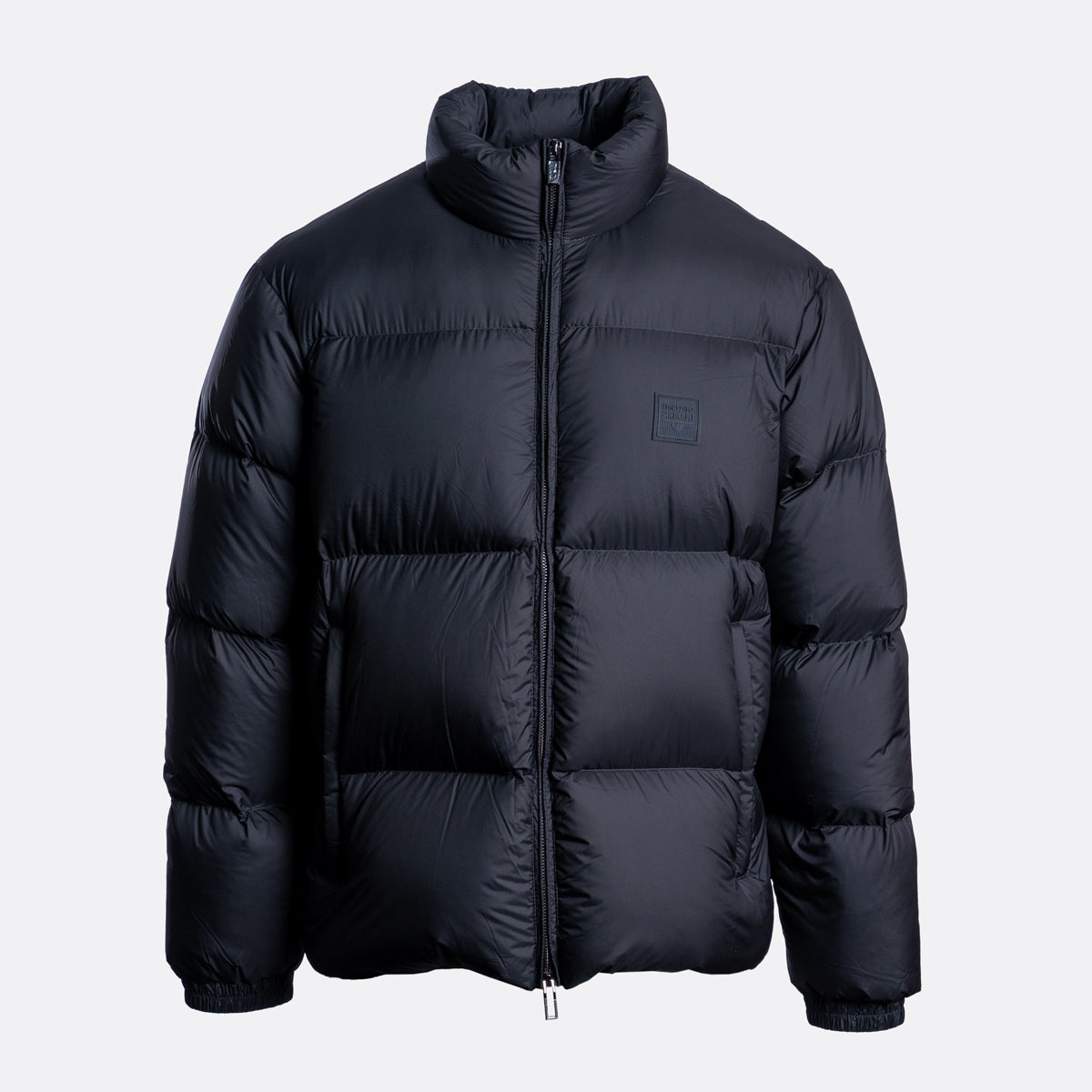 A black puffer jacket with horizontal quilting, a high stand-up collar, and a small tonal square logo patch on the left chest, shown against a light gray background. Emporio Armani Down Jacket. Sakks Menswear Southampton.