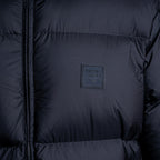 Close-up of a dark navy puffer jacket showing horizontal quilting, a front zipper, and a square black brand patch on the chest. Emporio Armani Down Jacket. Sakks Menswear Southampton.