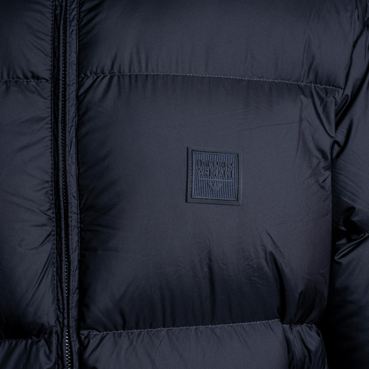 Close-up of a dark navy puffer jacket showing horizontal quilting, a front zipper, and a square black brand patch on the chest. Emporio Armani Down Jacket. Sakks Menswear Southampton.