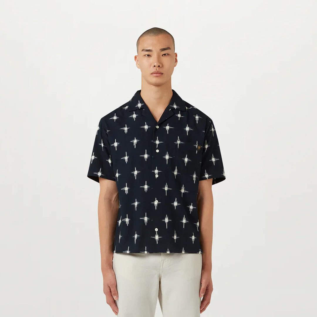 A man wearing a navy blue short-sleeve button-up shirt with a white geometric starburst pattern, paired with off-white trousers, standing against a plain white studio background. Belstaff Short Sleeve Shirt. Sakks Menswear Southampton.