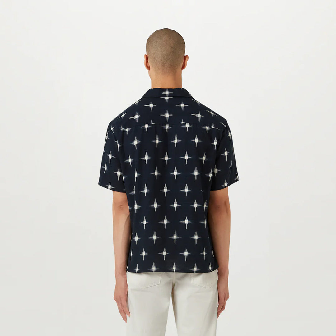 A man viewed from behind wearing a short-sleeve button-up shirt with a white geometric starburst pattern on a dark navy blue base, paired with off-white trousers, against a plain white studio background. Belstaff Short Sleeve Shirt. Sakks Menswear Southampton.