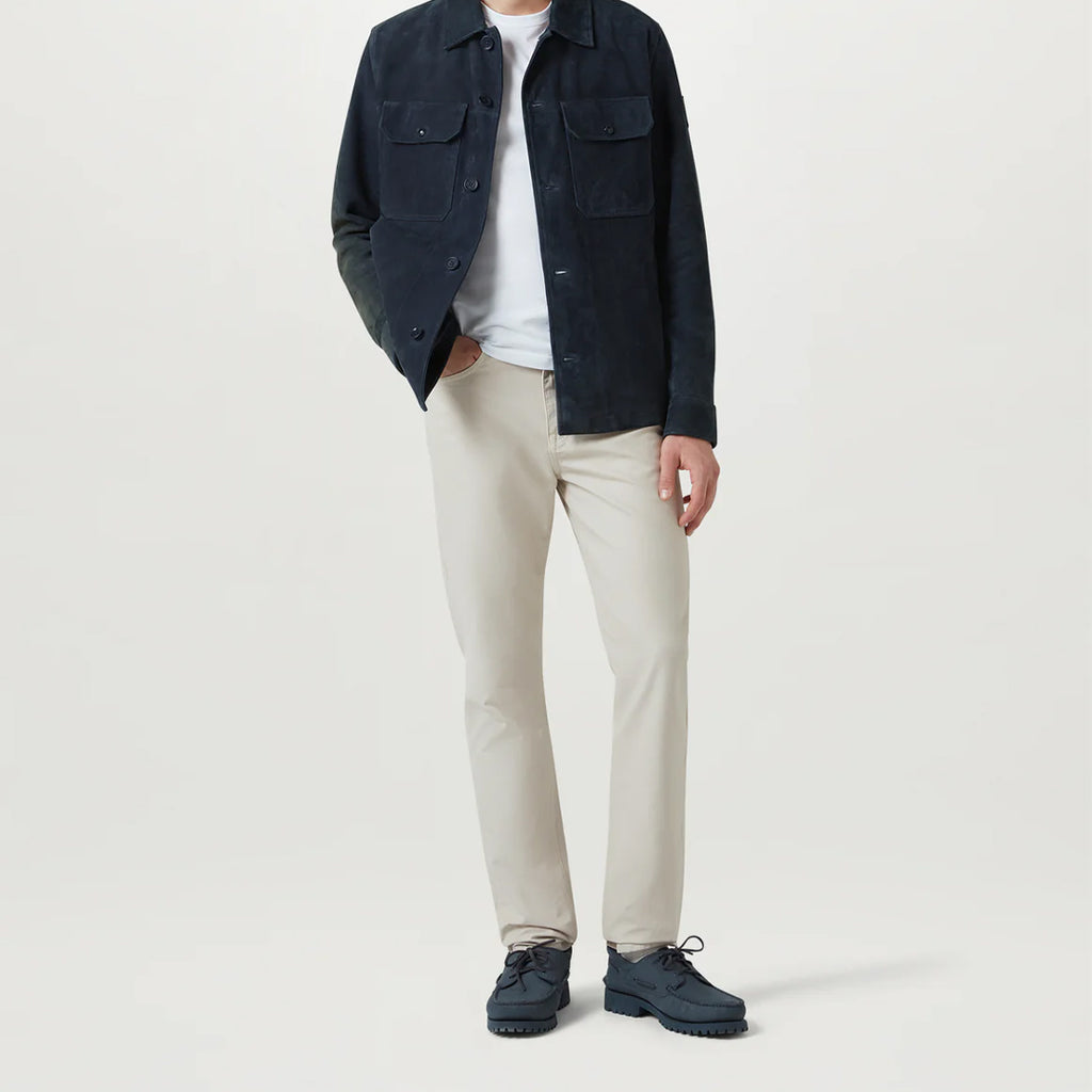 A model wearing a dark navy corduroy jacket over a white t-shirt, paired with beige straight-leg chinos and navy lace-up shoes, shot against a light gray studio background. Belstaff Chino available at Sakks Menswear in Southampton.