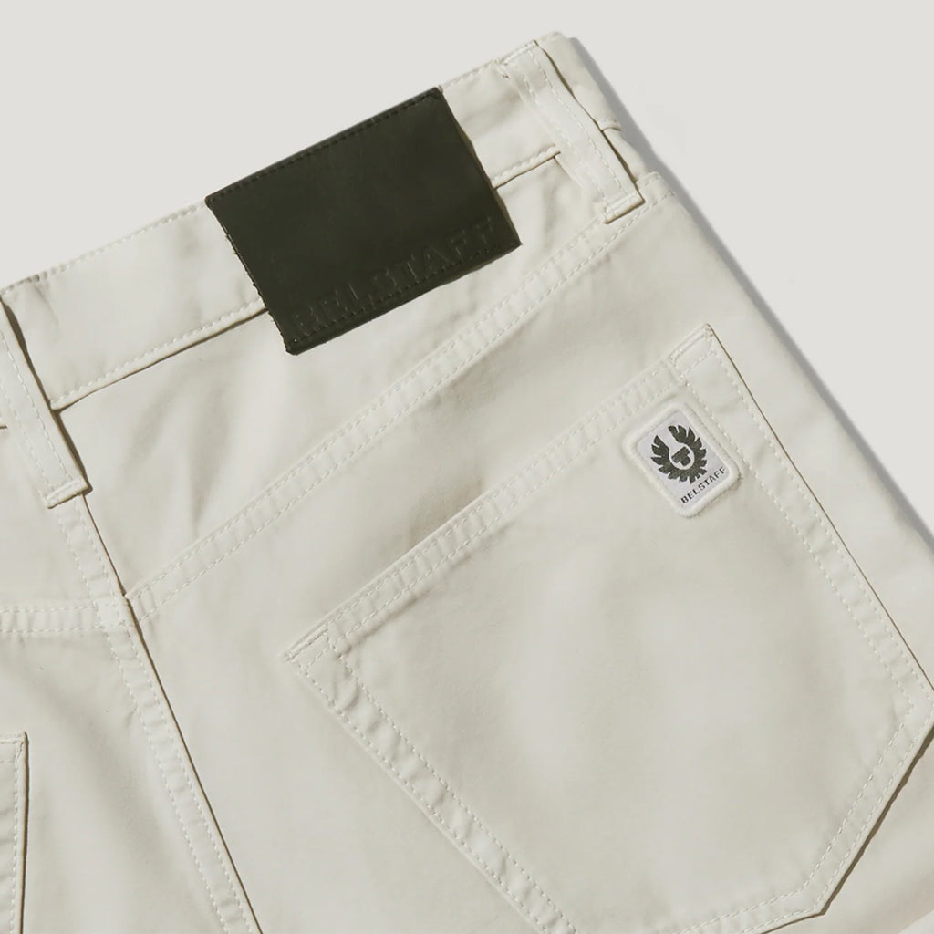 Close-up of the back waistband and pocket area of light beige denim jeans, showing a dark olive brand patch at the back waist and a small white embroidered Paddyus logo patch on the pocket. Belstaff Chino available at Sakks Menswear in Southampton.