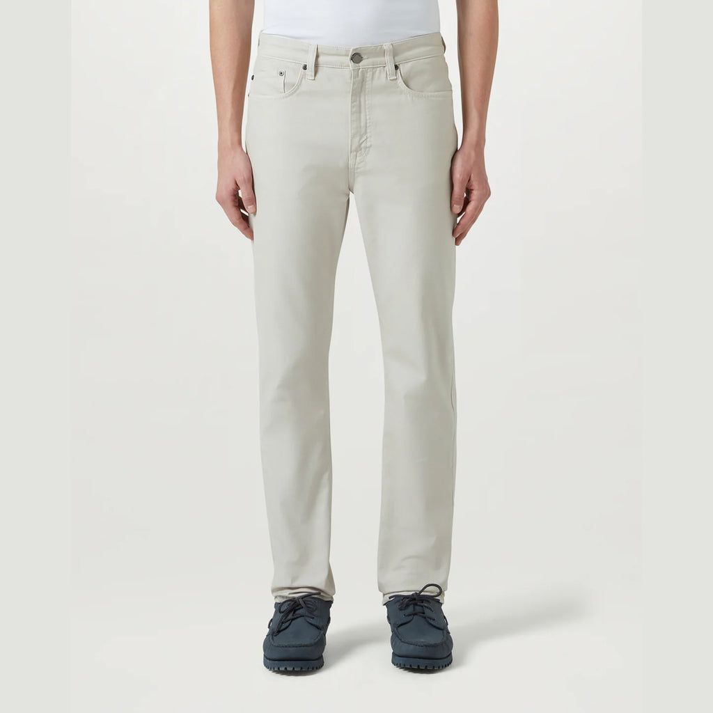 A person wearing light beige straight-leg jeans with a white top and dark blue shoes, standing against a plain light gray studio background. Belstaff Chino available at Sakks Menswear in Southampton.