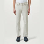 A person wearing light beige straight-leg jeans with a white top and dark blue shoes, standing against a plain light gray studio background. Belstaff Chino available at Sakks Menswear in Southampton.