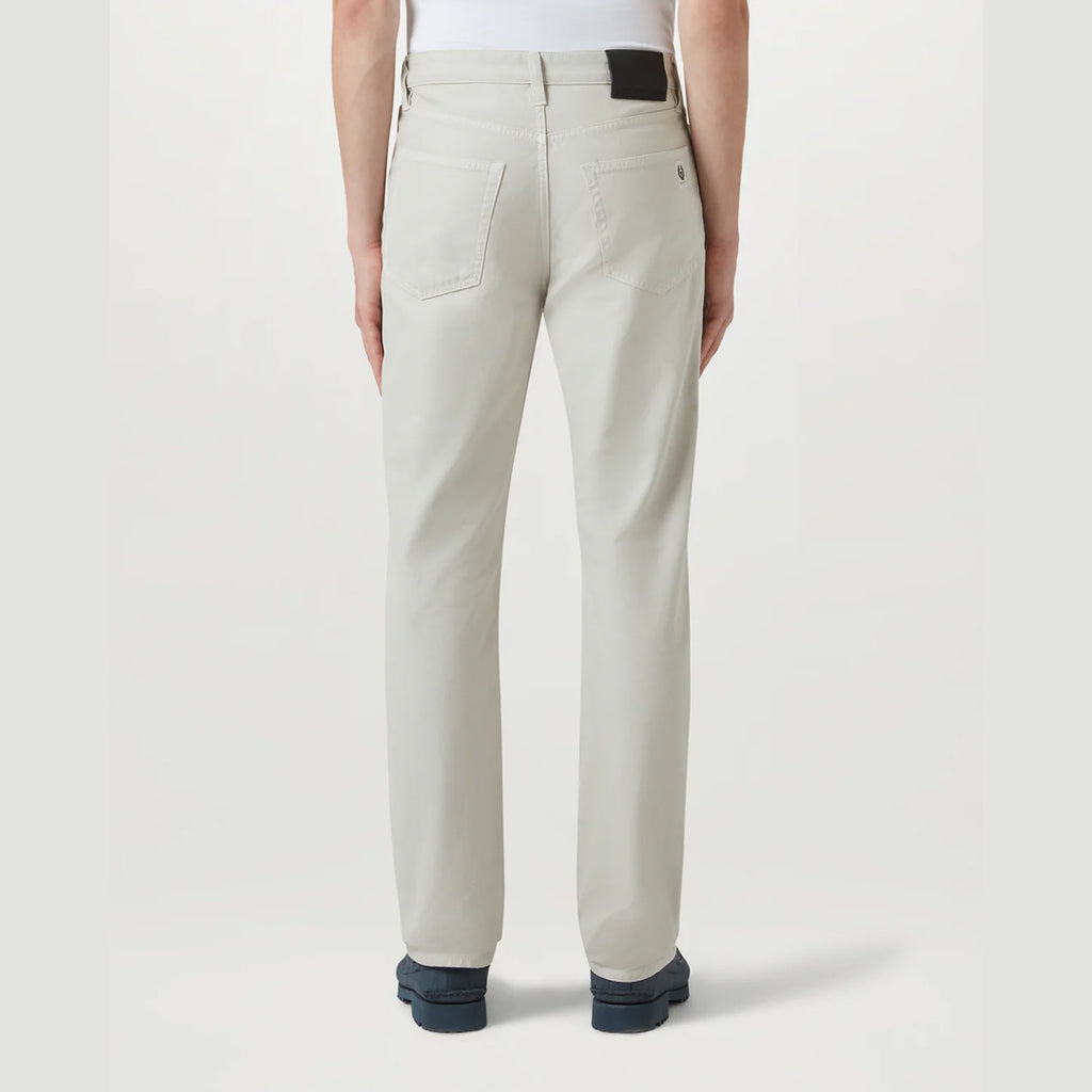 A person shown from the back wearing light beige straight-leg jeans, a white top, and dark blue shoes against a light gray studio background. Belstaff Chino available at Sakks Menswear in Southampton.