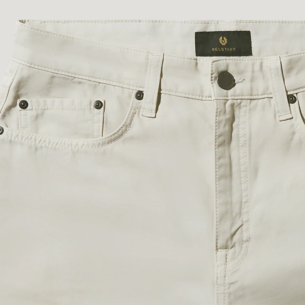 Close-up of a pair of light beige denim jeans showing the waistband, belt loops, front button closure, and pocket details. A black BELSTAFF label is visible at the top center. Belstaff Chino available at Sakks Menswear in Southampton.