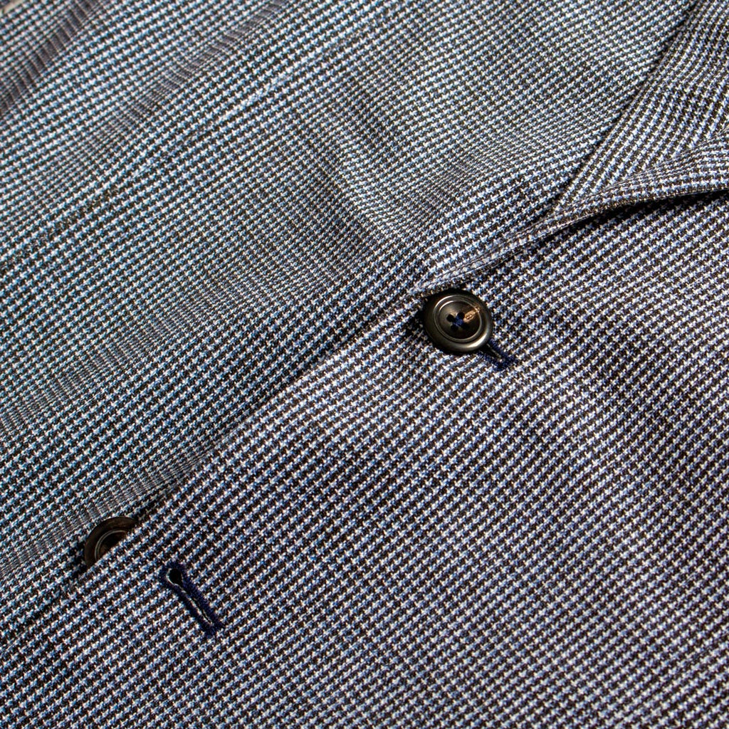 Close-up of a textured woven fabric with a fine micro-check pattern in gray, white, and subtle blue tones, featuring a dark metal button and a small dark accent. Circolo 1901 Blazer available at Sakks Menswear in Southampton.
