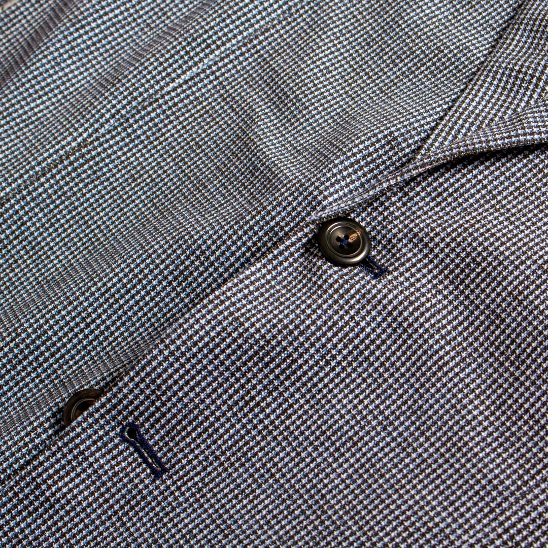 Close-up of a textured woven fabric with a fine micro-check pattern in gray, white, and subtle blue tones, featuring a dark metal button and a small dark accent. Circolo 1901 Blazer available at Sakks Menswear in Southampton.