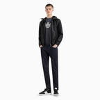 A male model stands against a plain light gray studio background wearing a black hooded jacket over a black graphic T-shirt, dark navy pants, and black-and-white sneakers. Emporio Armani Jeans. Sakks Menswear Southampton.