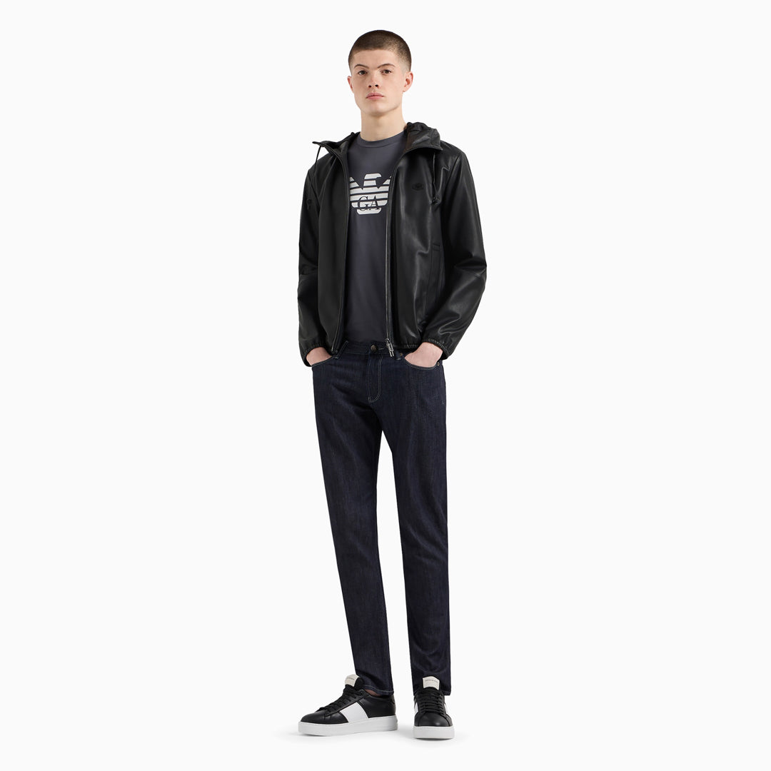 A male model stands against a plain light gray studio background wearing a black hooded jacket over a black graphic T-shirt, dark navy pants, and black-and-white sneakers. Emporio Armani Jeans. Sakks Menswear Southampton.