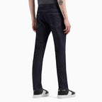 Rear view of a person wearing dark indigo slim-fit jeans with black and white sneakers, shown against a plain light gray studio background. Emporio Armani Jeans. Sakks Menswear Southampton.