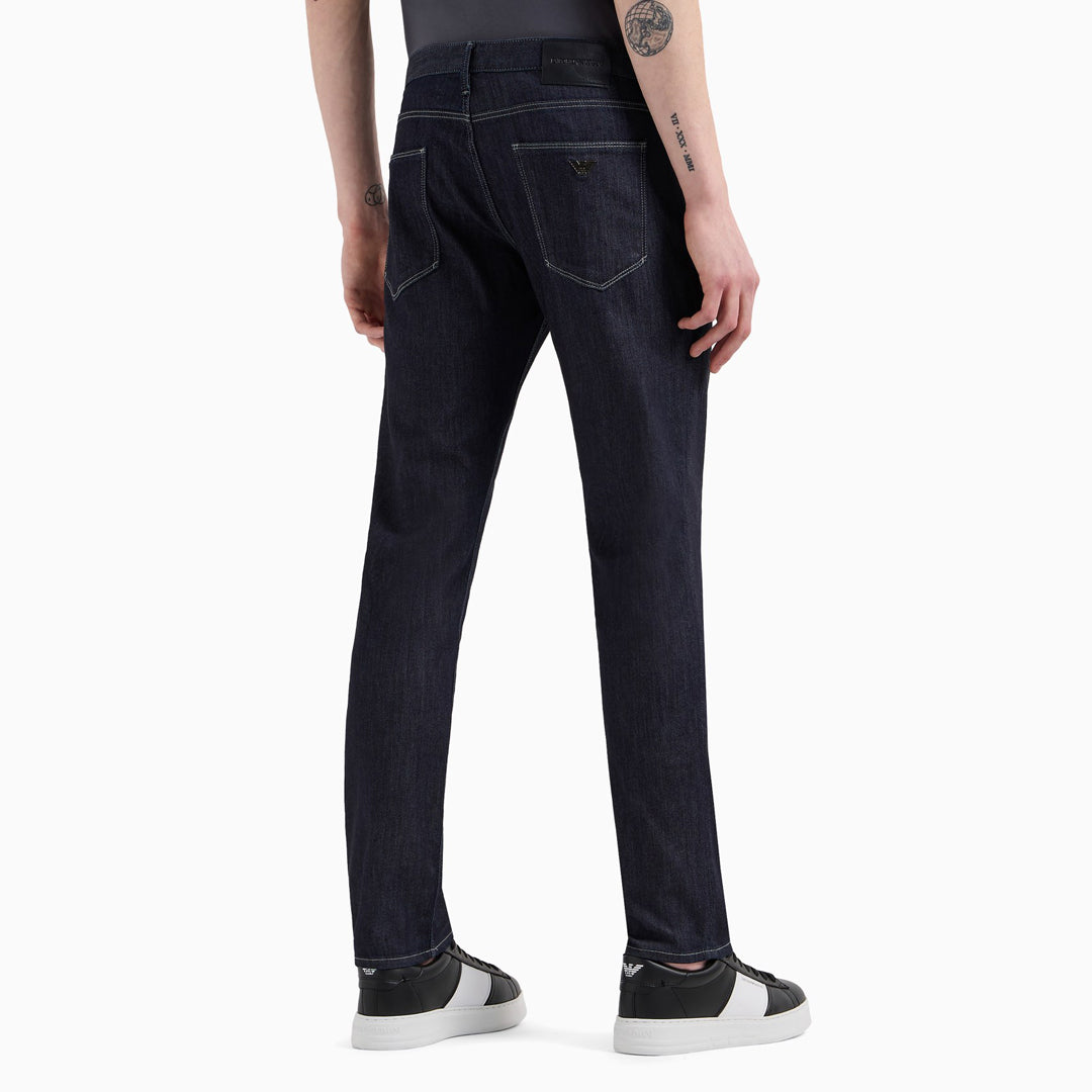 Rear view of a person wearing dark indigo slim-fit jeans with black and white sneakers, shown against a plain light gray studio background. Emporio Armani Jeans. Sakks Menswear Southampton.