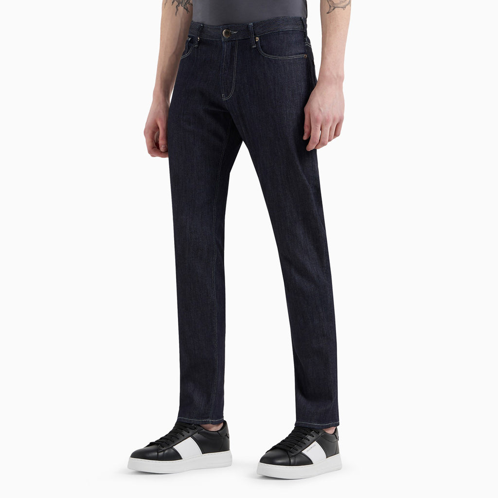 A person wearing dark indigo straight-leg jeans with a gray t-shirt and black-and-white sneakers, shown from the waist down on a light gray studio background. Emporio Armani Jeans. Sakks Menswear Southampton.