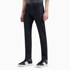 A person wearing dark indigo straight-leg jeans with a gray t-shirt and black-and-white sneakers, shown from the waist down on a light gray studio background. Emporio Armani Jeans. Sakks Menswear Southampton.