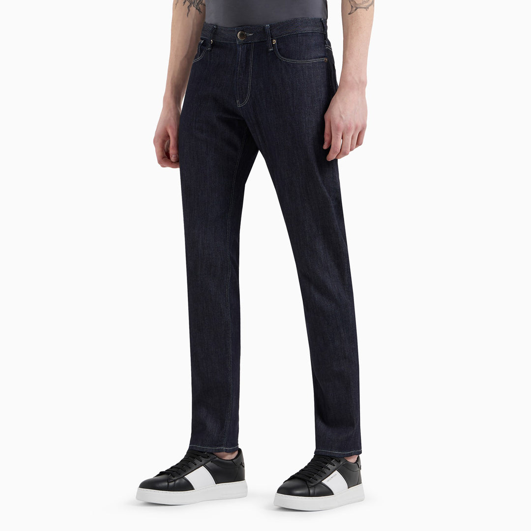 A person wearing dark indigo straight-leg jeans with a gray t-shirt and black-and-white sneakers, shown from the waist down on a light gray studio background. Emporio Armani Jeans. Sakks Menswear Southampton.