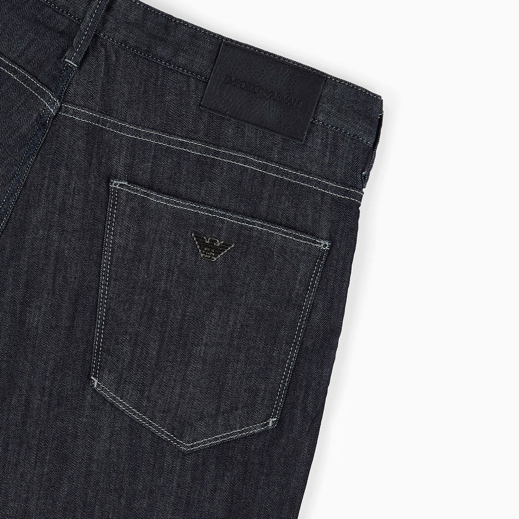Close-up of the back waistband and pocket area of dark indigo denim jeans, showing a black woven brand patch and a small metallic eagle logo on the pocket. Emporio Armani Jeans. Sakks Menswear Southampton.