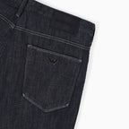 Close-up of the back waistband and pocket area of dark indigo denim jeans, showing a black woven brand patch and a small metallic eagle logo on the pocket. Emporio Armani Jeans. Sakks Menswear Southampton.