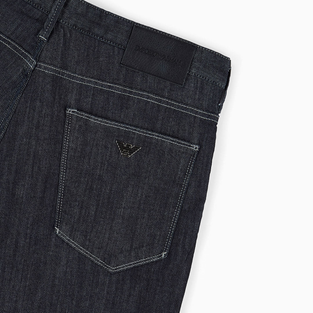 Close-up of the back waistband and pocket area of dark indigo denim jeans, showing a black woven brand patch and a small metallic eagle logo on the pocket. Emporio Armani Jeans. Sakks Menswear Southampton.