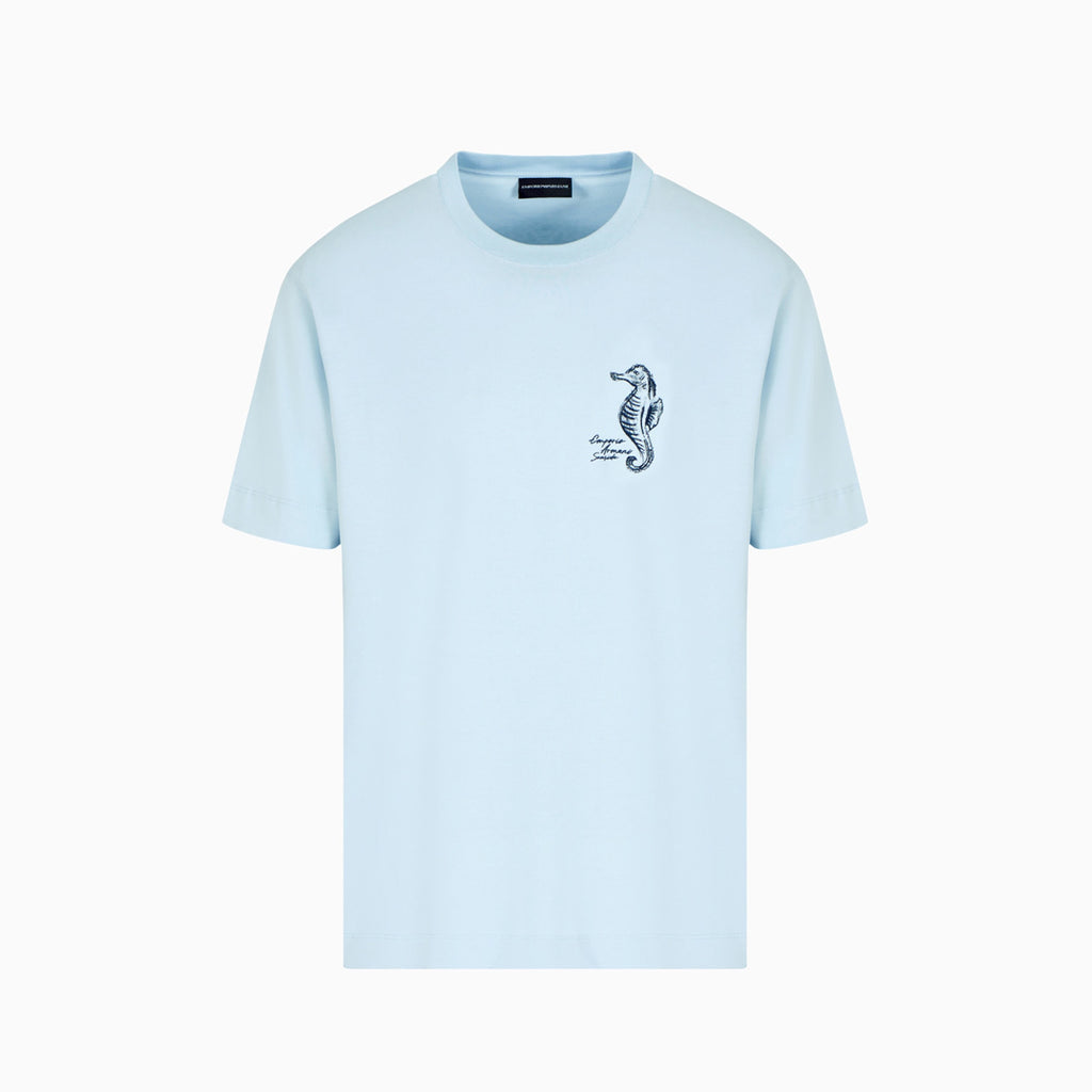 A light blue short-sleeve crewneck t-shirt with a small black seahorse graphic on the left chest, displayed on a clean white background. Emporio Armani T-Shirt. Sakks Menswear Southampton.
