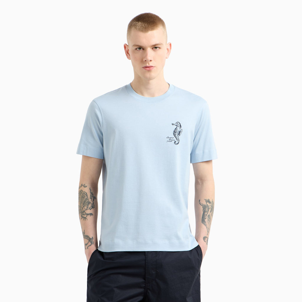 A man wearing a light blue crewneck t-shirt with a small black seahorse graphic on the left chest, paired with dark navy pants, set against a plain light gray background. Emporio Armani T-Shirt. Sakks Menswear Southampton.