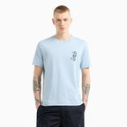 A man wearing a light blue crewneck t-shirt with a small black seahorse graphic on the left chest, paired with dark navy pants, set against a plain light gray background. Emporio Armani T-Shirt. Sakks Menswear Southampton.