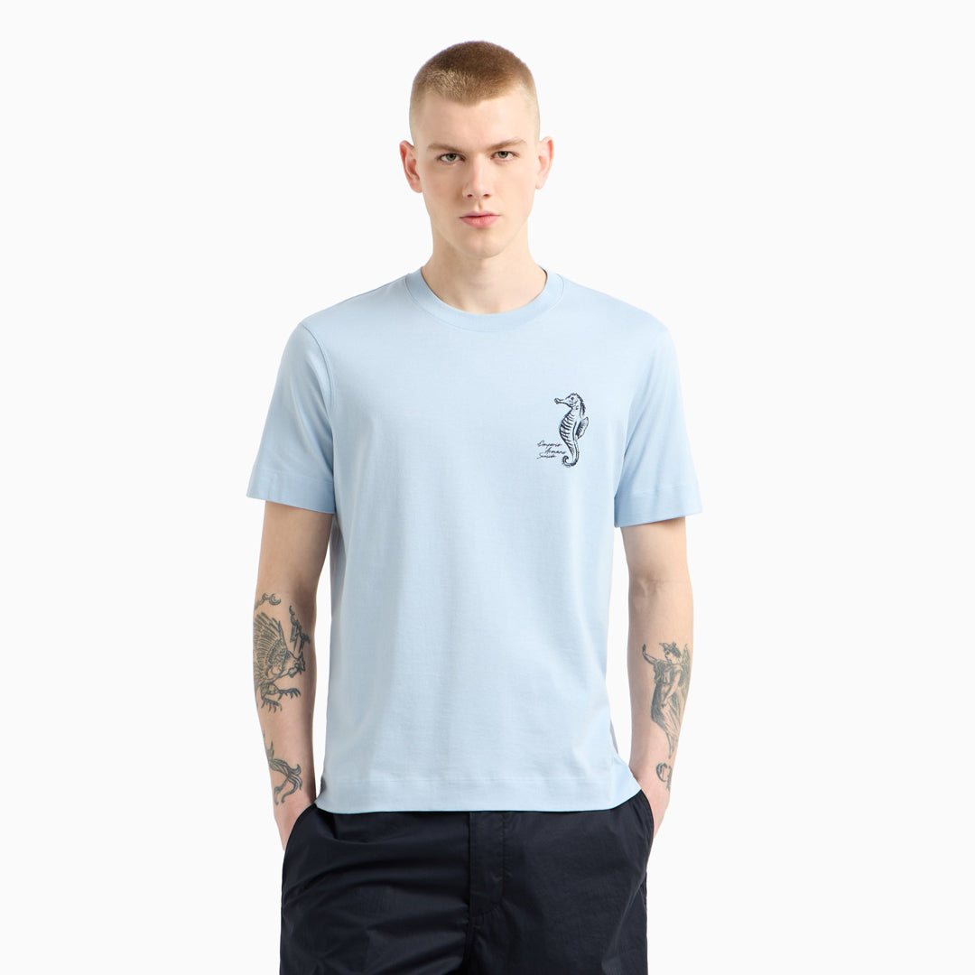A man wearing a light blue crewneck t-shirt with a small black seahorse graphic on the left chest, paired with dark navy pants, set against a plain light gray background. Emporio Armani T-Shirt. Sakks Menswear Southampton.