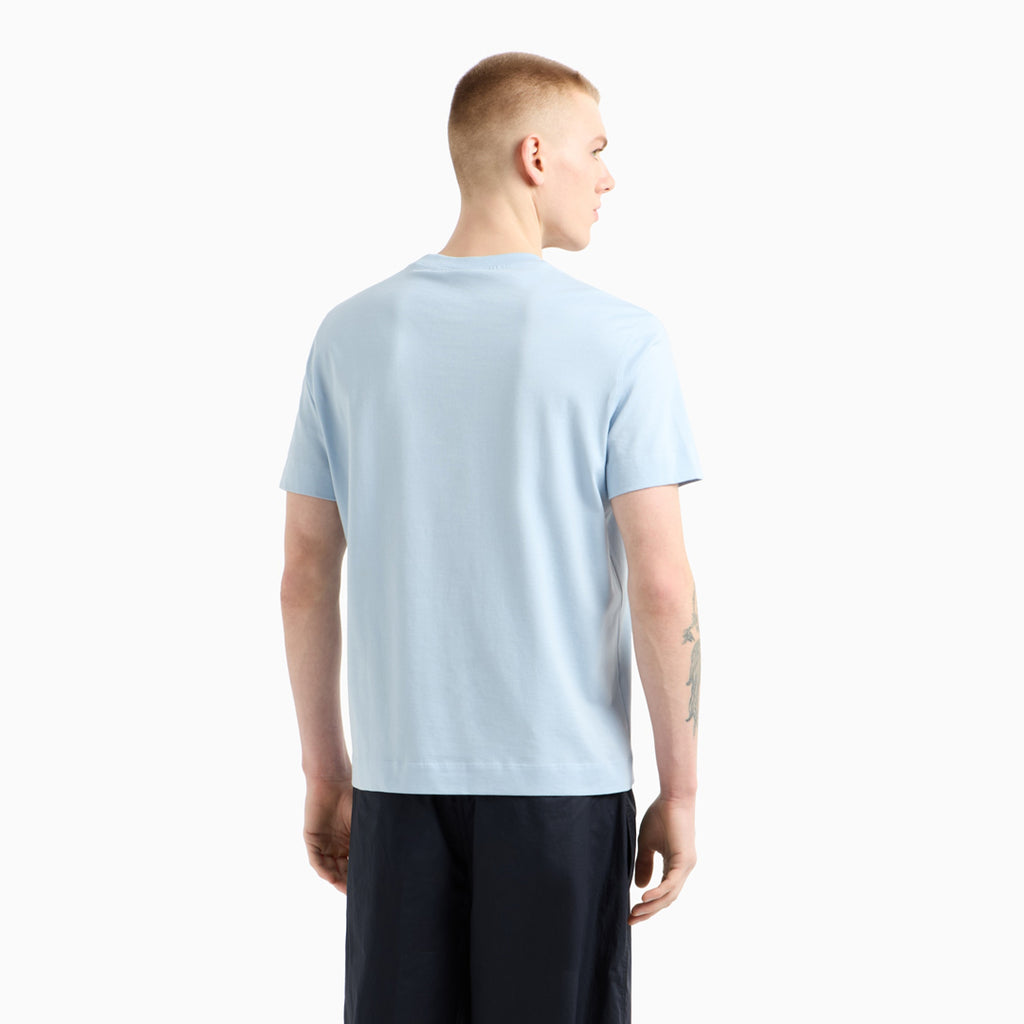 A man viewed from the back wearing a light blue crewneck t-shirt and dark navy pants, set against a plain white studio background. Emporio Armani T-Shirt. Sakks Menswear Southampton.