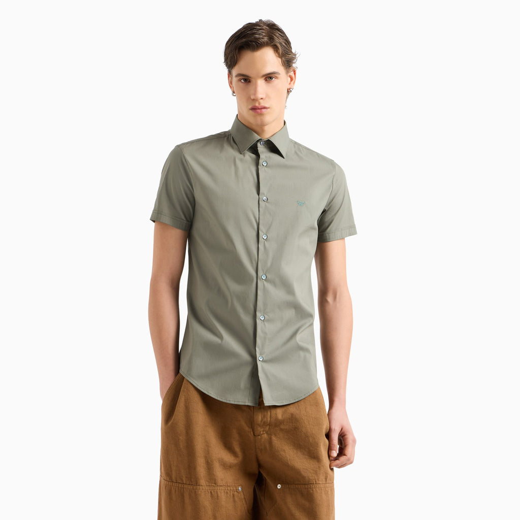 A man modeling a short-sleeve button-up shirt in sage green, paired with brown pants, against a plain white background. Emporio Armani Short Sleeved Shirt. Sakks Menswear Southampton.