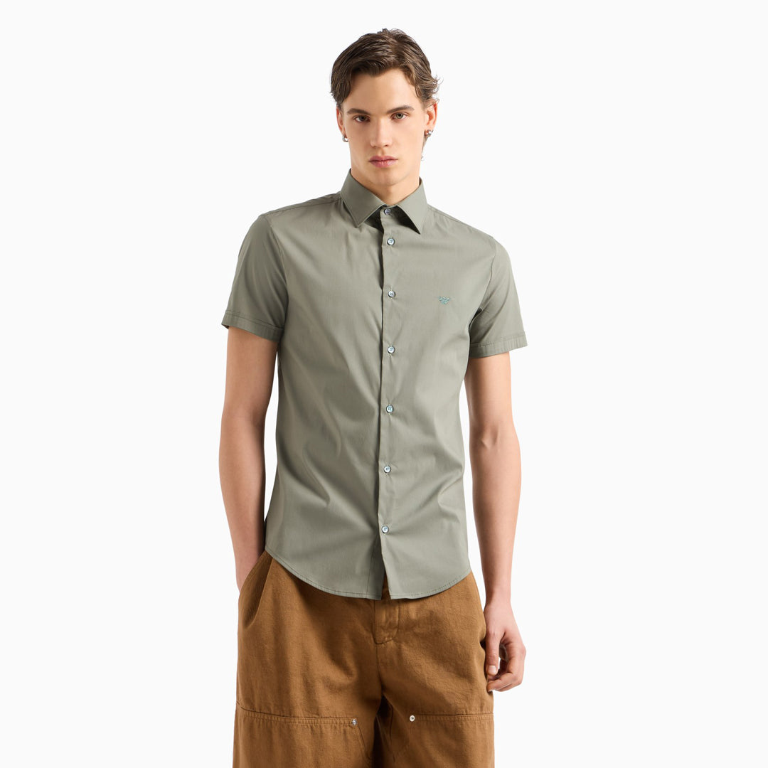 A man modeling a short-sleeve button-up shirt in sage green, paired with brown pants, against a plain white background. Emporio Armani Short Sleeved Shirt. Sakks Menswear Southampton.