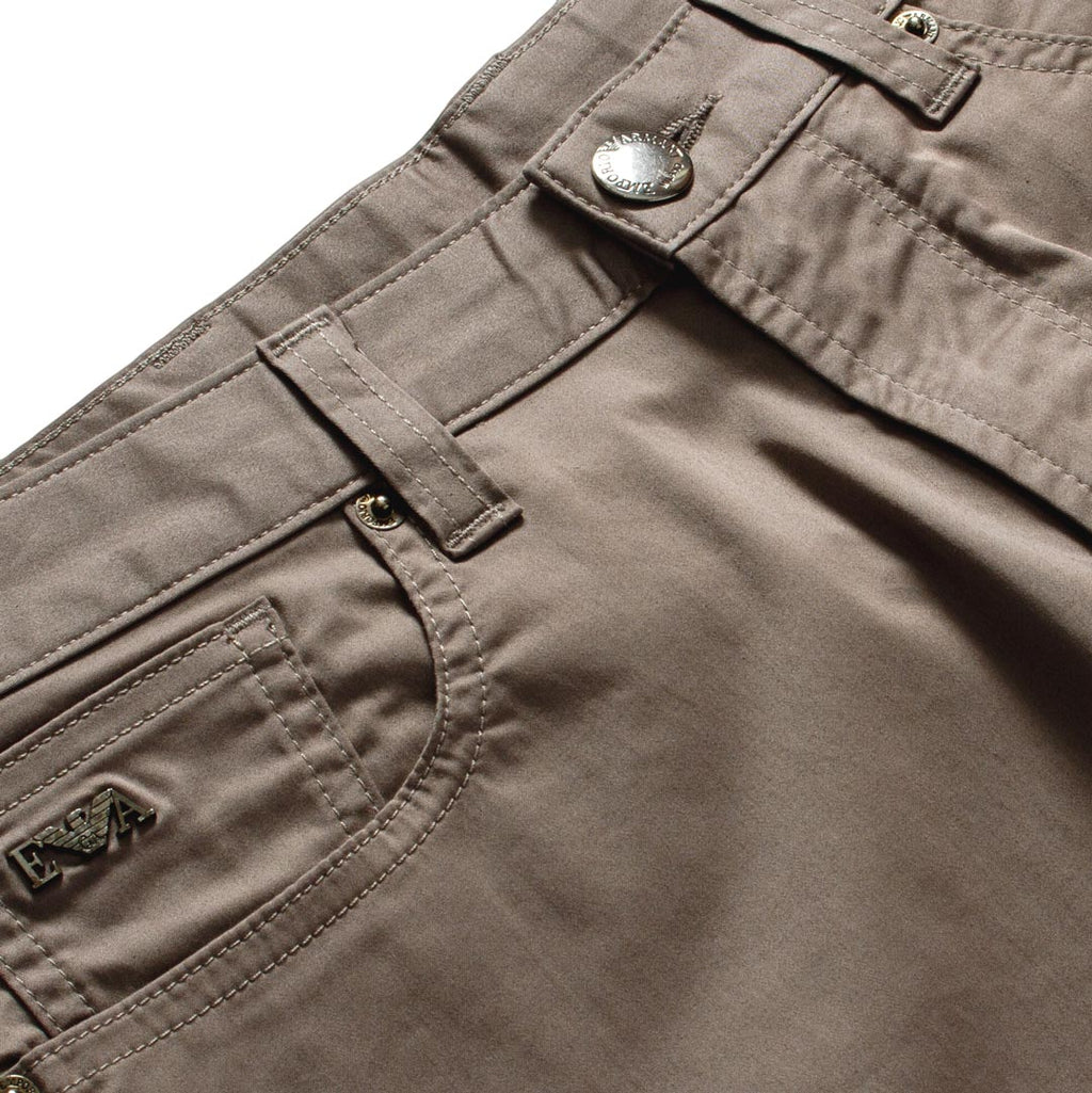 Close-up of the waistband and front pocket of taupe brown pants, showing a silver metal button, belt loops, and a small embroidered logo patch on the pocket. Emporio Armani Shorts. Sakks Menswear Southampton.