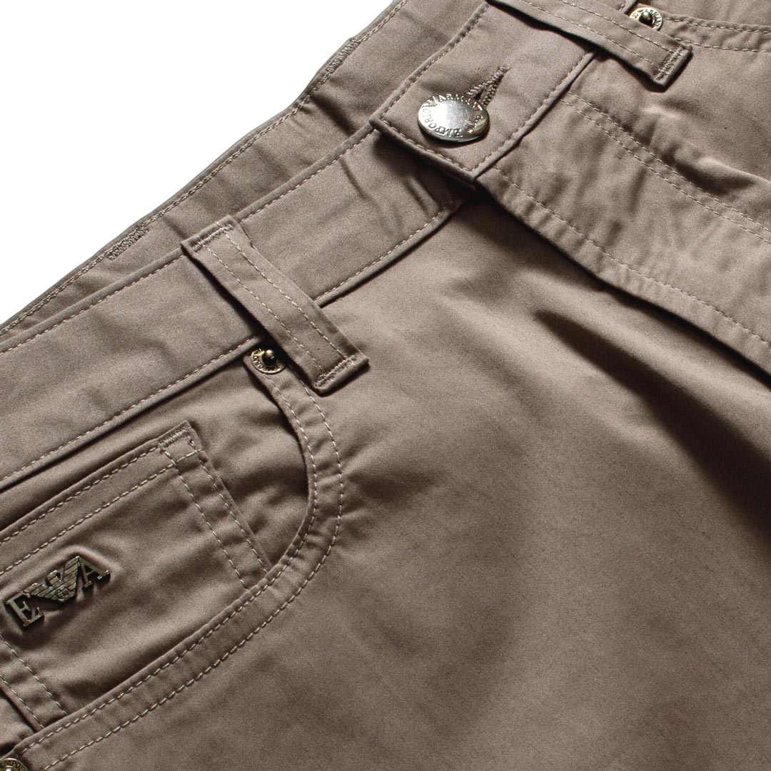 Close-up of the waistband and front pocket of taupe brown pants, showing a silver metal button, belt loops, and a small embroidered logo patch on the pocket. Emporio Armani Shorts. Sakks Menswear Southampton.