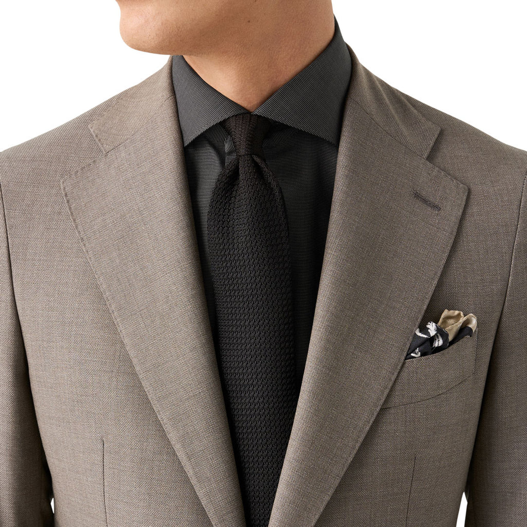 A close-up of a man wearing a light brown textured suit jacket, a dark charcoal dress shirt, a black textured tie, and a patterned pocket square in his breast pocket, set against a plain white background. Eton Shirt available at Sakks Menswear in Southampton.