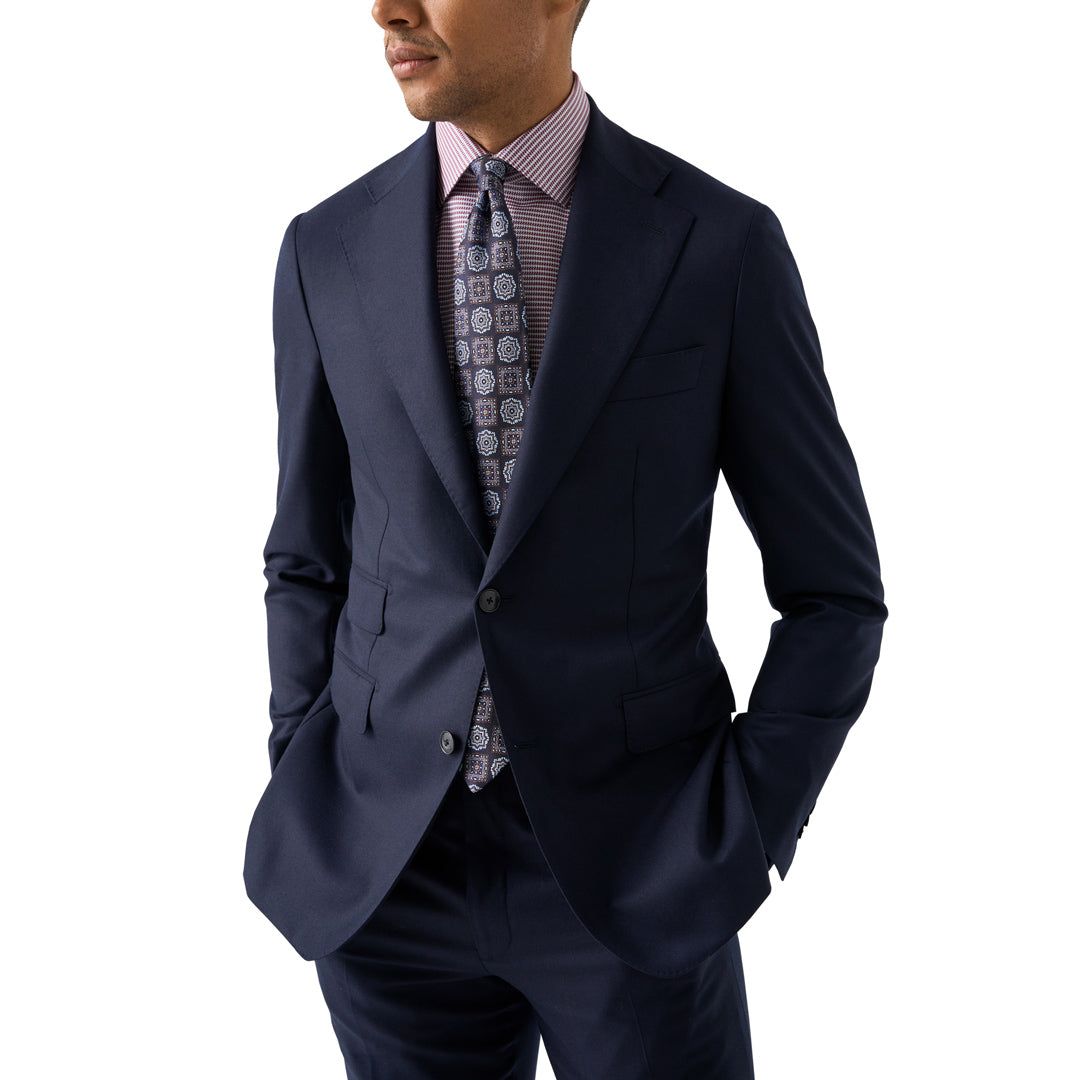 A man wearing a navy blue suit jacket and matching trousers, styled with a pink patterned dress shirt and a patterned tie, against a solid white background. Eton Shirt available at Sakks Menswear in Southampton.