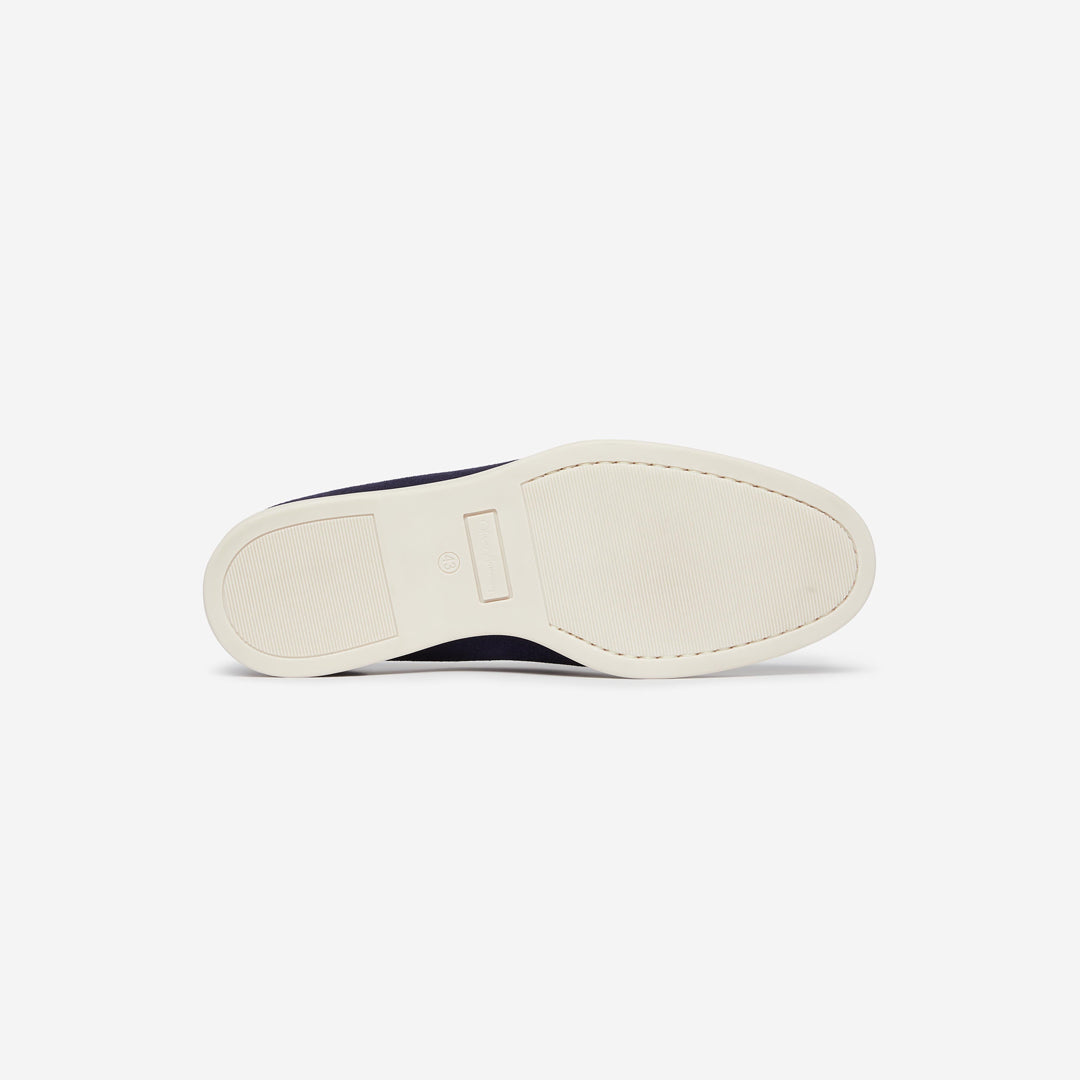 The outsole view of a slip-on shoe, showing a thick cream-colored rubber sole with a dark navy blue upper peeking out from beneath, set against a plain light gray background. Oliver Sweeney Loafers available at Sakks Menswear in Southampton.