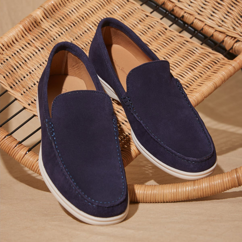 A pair of navy blue suede slip-on loafers with white soles, displayed on a woven wicker chair over a light wood floor. Oliver Sweeney Loafers available at Sakks Menswear in Southampton.