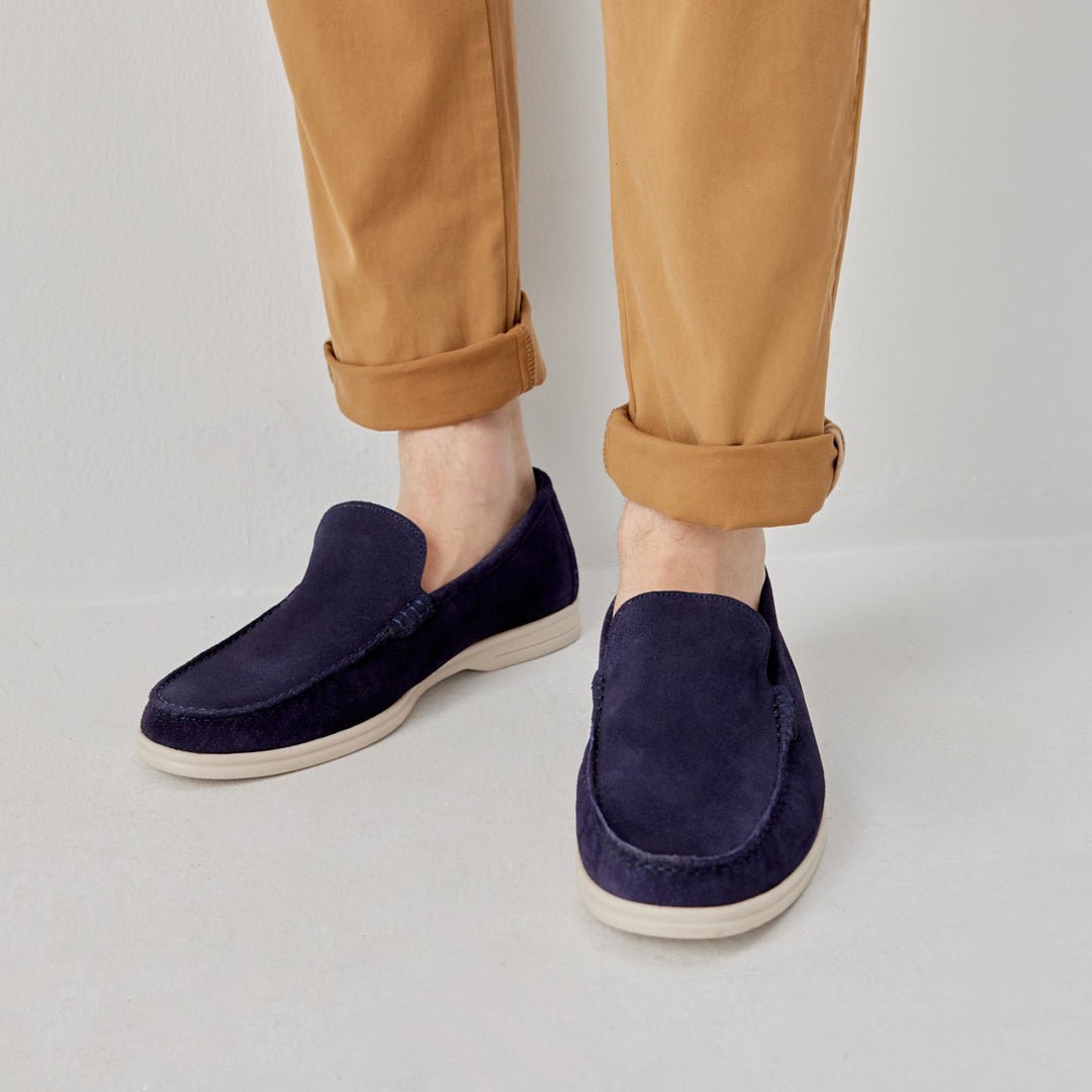 A person wearing navy blue suede slip-on loafers with off-white soles, paired with tan cuffed trousers, shown from the calves down against a light gray studio background. Oliver Sweeney Loafers available at Sakks Menswear in Southampton.