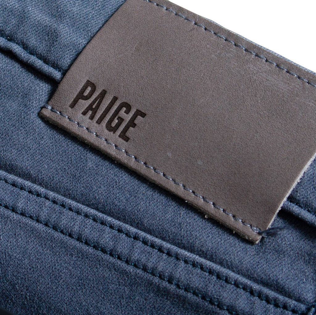 Close-up of a pair of blue denim jeans showing the back pocket with a brown leather patch embossed with the brand name 'PAIGE'. PAIGE Jeans available at Sakks Menswear in Southampton.