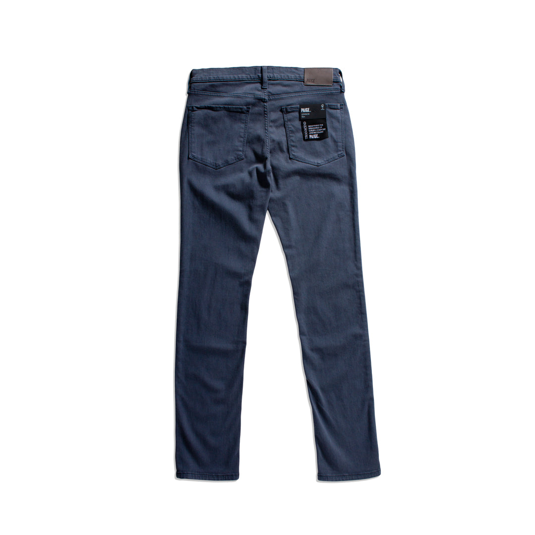 Back view of a pair of dark blue denim jeans with a brown leather patch on the waistband and a black brand tag on the back pocket, shown on a plain white background. PAIGE Jeans available at Sakks Menswear in Southampton.