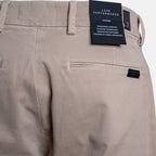 Close-up of the back waistband of beige chino pants, showing belt loops, a rear welt pocket, and a black hangtag labeled Luxe Performance. - 7 For All Mankind Comfort Sateen Chino Short in Mud. Sakks Menswear.