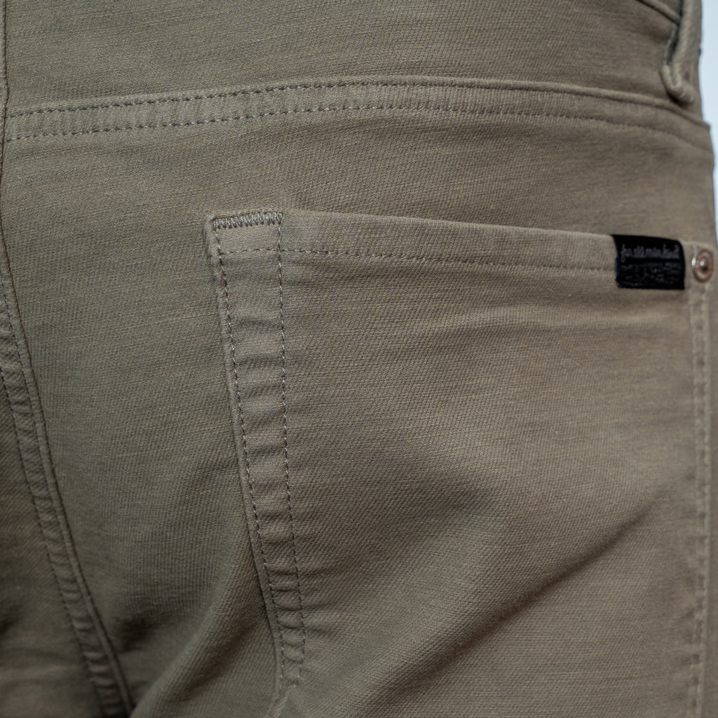 Close-up of the back pocket of a pair of khaki-colored denim jeans, showing the stitching and a small black brand label. 7 For All Mankind. Sakks Menswear Southampton.
