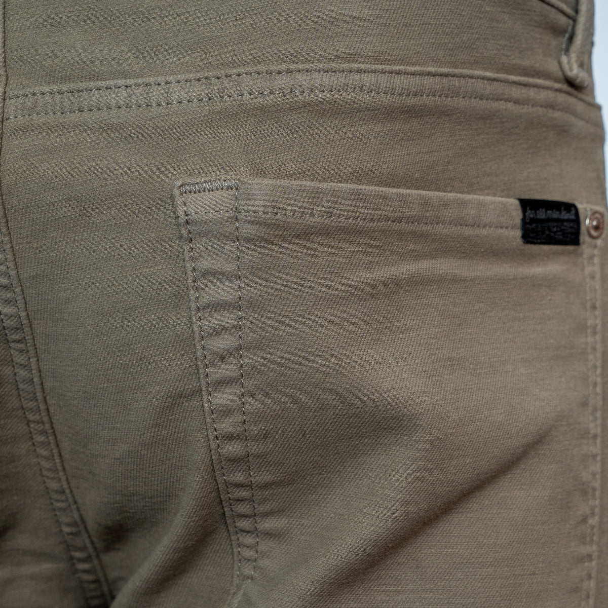 Close-up of the back pocket of a pair of khaki-colored denim jeans, showing the stitching and a small black brand label. 7 For All Mankind. Sakks Menswear Southampton.