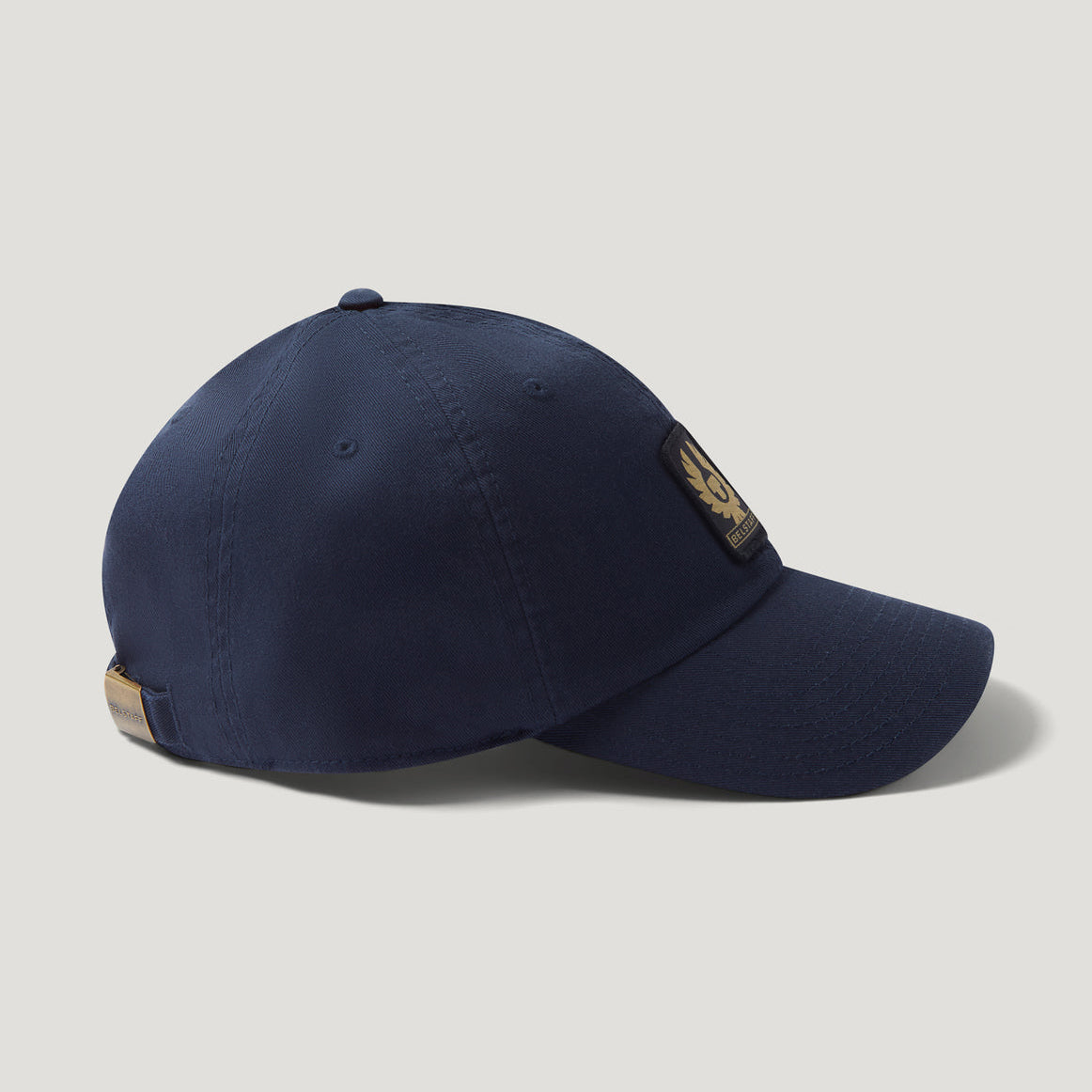 A navy blue baseball cap with a curved brim, featuring a gold embroidered winged logo patch on the side and a tan leather tag on the back, set against a light gray background. Belstaff Cap. Sakks Menswear Southampton.