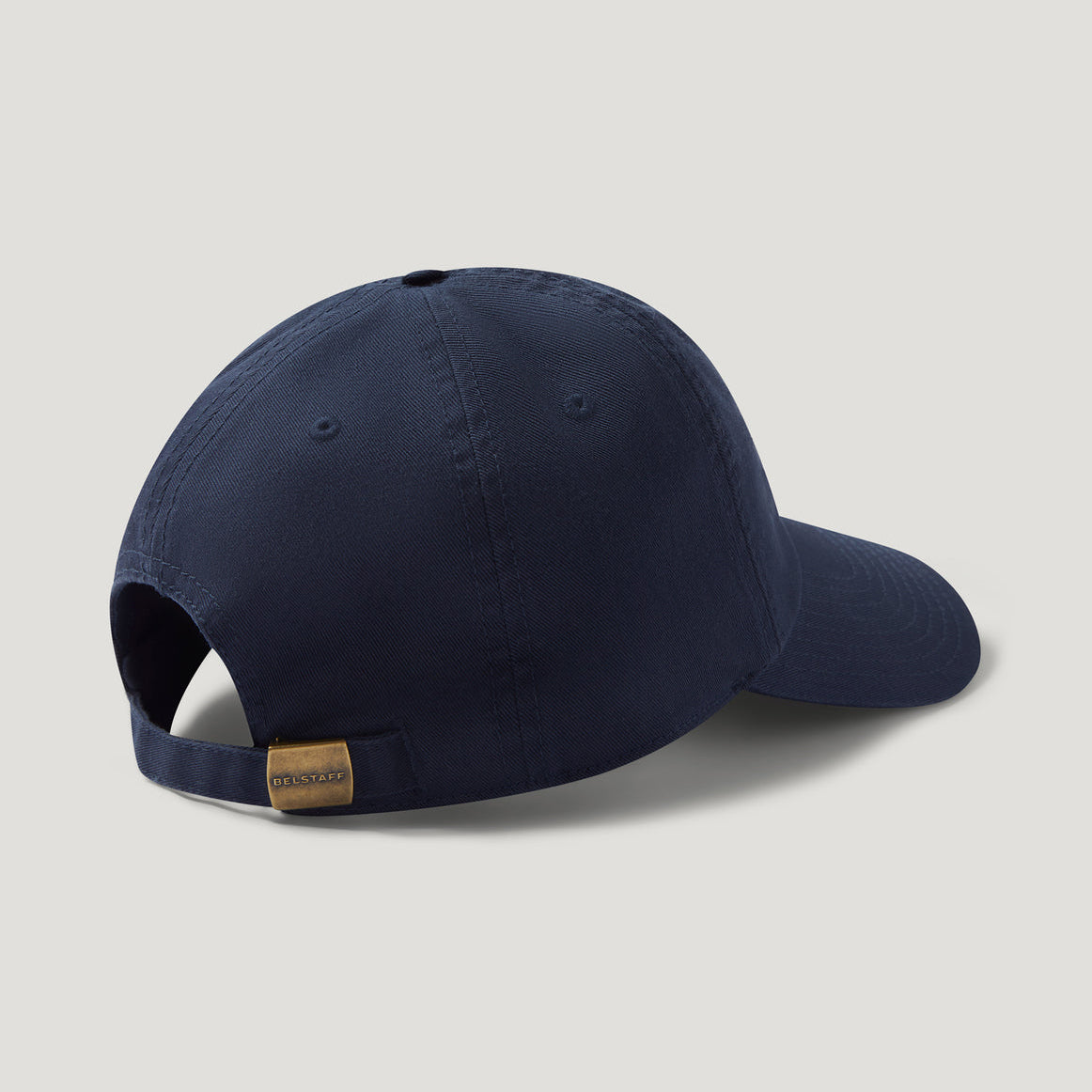 A navy blue baseball cap shown from a rear three-quarter angle, featuring a small tan leather brand tab on the back strap, set against a light gray background. Belstaff Cap. Sakks Menswear Southampton.