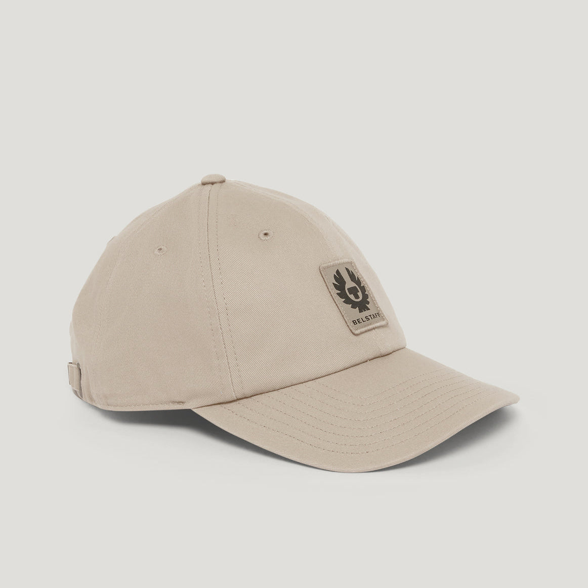 A light beige baseball cap with a curved brim and a rectangular Barley patch on the front, displayed on a neutral light gray background. Belstaff Cap. Sakks Menswear Southampton.