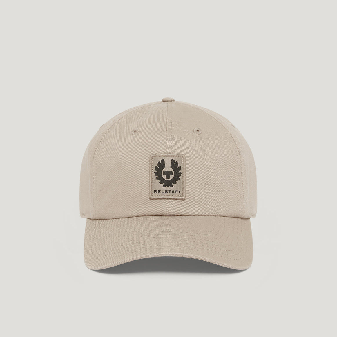 A light beige baseball cap with a curved brim and a rectangular Barley patch on the front, displayed on a neutral light gray background. Belstaff Cap. Sakks Menswear Southampton.